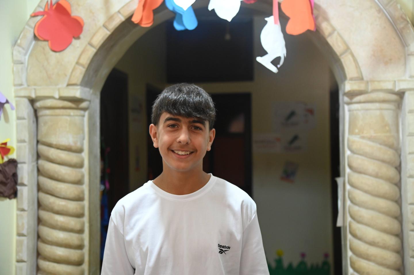 Alaa, 15, smiles while standing inside a learning centre in Wadi Khaled, Akkar, surrounded by colourful handmade decorations.