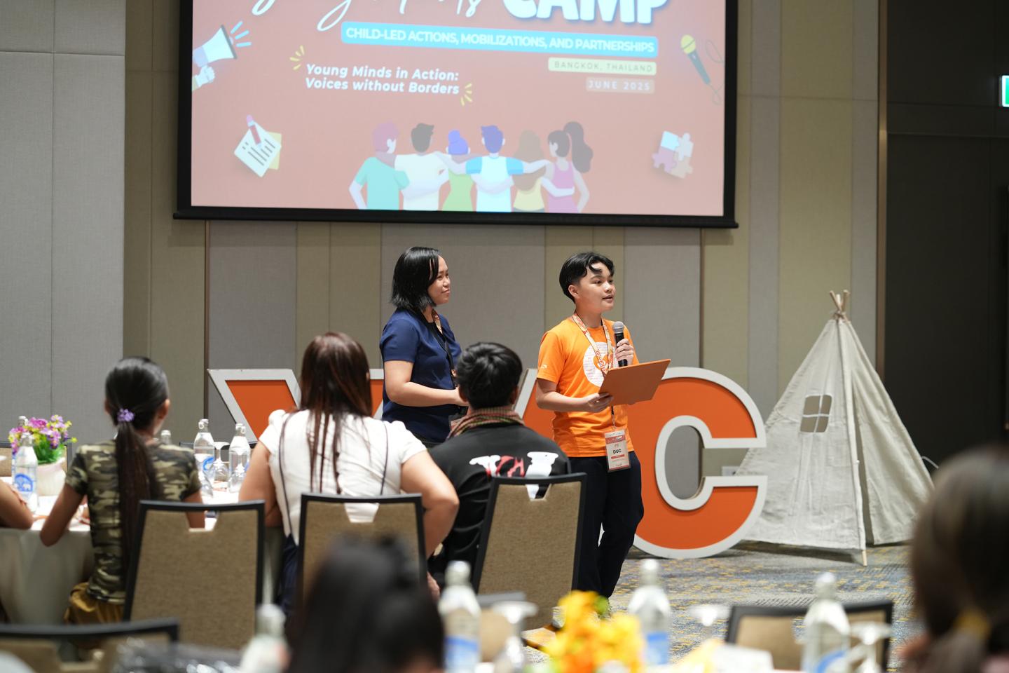 A youth stands in front of a room of other youth holding a clipboard and speaking into a microphone.