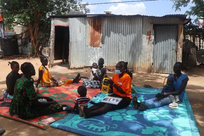 A group of women with their babies sit on the ground listening to a radio