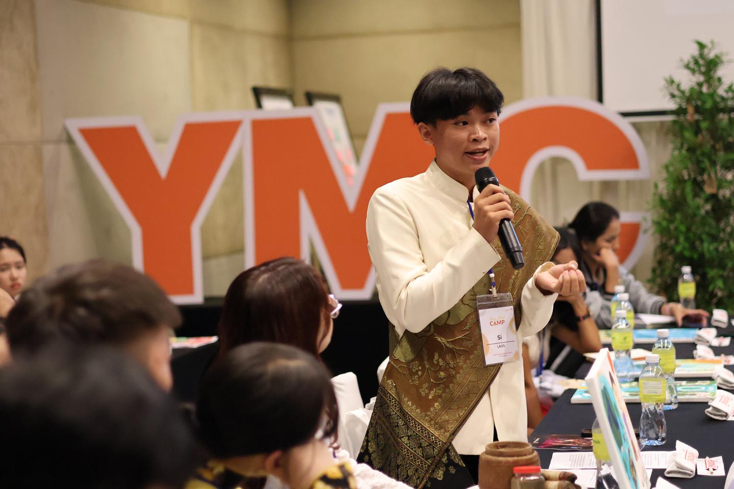 See, a youth from Cambodia, stands at a table speaking into a microphone.