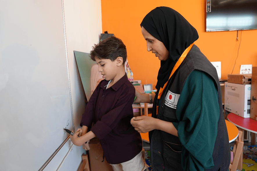 Abdallah* writing on the board with the support of the assistant teacher. World Vision Syria Response, Joanna Zreineh.