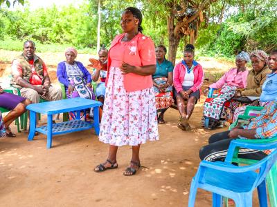 Felister Charles Mbavani (center), a farmer and Trainer of Trainers (ToT), shares her story of transformation at her home in Nganwa Village, Makueni County. Seated around her are members of the Lovers Self-Help Group, gathered for their weekly Village Saving and Loan Association (VSLA) table banking meeting. © World Vision Kenya/ Jared Ontobo