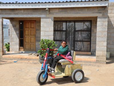 Reithabetse, a 7-year-old girl from Lesotho, smiling outside her home while sitting on her new mobility cart.