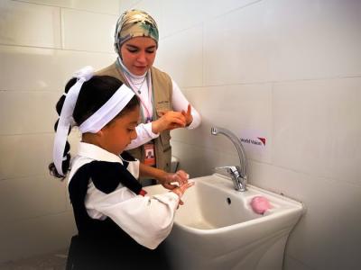 A young girl in a school uniform washes her hands with the help of a female World Vision staff member