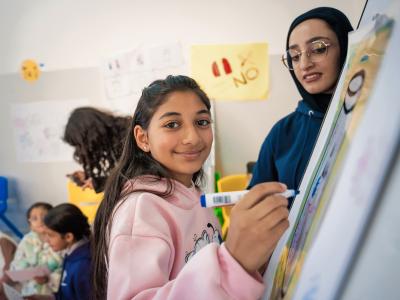 A young girl in the West Bank smiles at the camera while standing at a white board with marker in hand.
