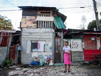 A girl in a white shirt and pink skirt stands in front of an urban settlement