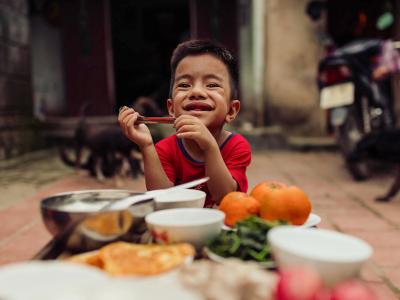 A young Vietnamese boy smile to the camera. He is holding a spoon with bowls and plates of food on a table in front of him. 