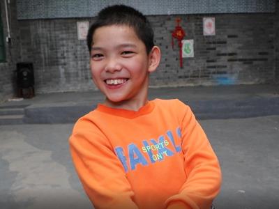A young Chinese boy with cerebral palsy wears an orange shirt and smiles at the camera