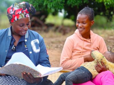 Two woman sitting on a grass mat, one breast-feeding a baby.