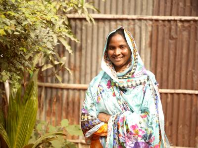 Dinajpur community leader Sufia smiling outside her home, representing empowerment and local leadership in Bangladesh.