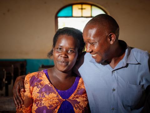 A husband and wife stand smiling together in front of a church stained glass window