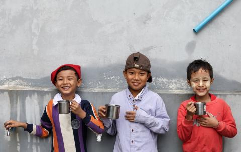 Three boys stand in front of a wall holding tin cups filled with water. One boy is also holding onto a water spout.