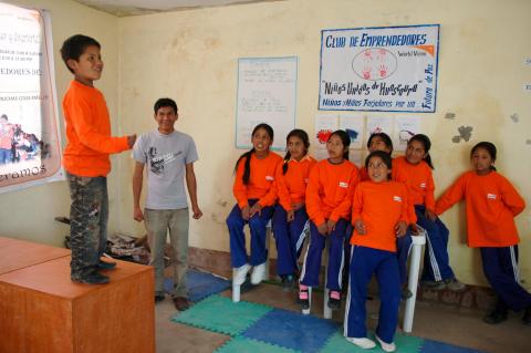 A boy in a long sleeve orang shirt, stands on a desk and talks to a group of children, also in orange long sleeve shirts