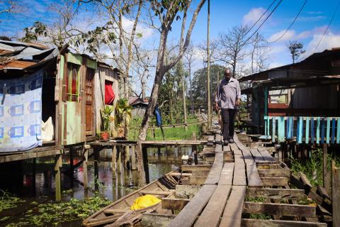 A man walks on wood planks over a body of water, surrounded by wood structures on stilts.