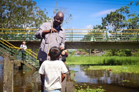 A man holds a fishing line with a small fish as a boy, with his back to the camera, holds the fishing pole.