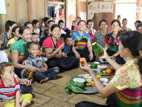 A group of women and children sit on the ground while looking at a woman holding vegetables.