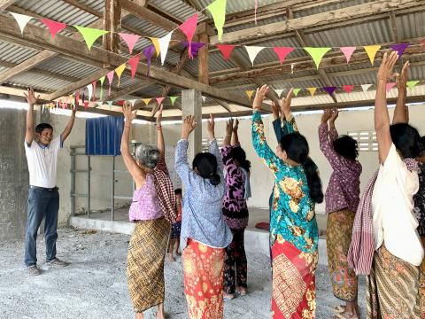 A community health worker stands with his arms raised in front of a group of women who alos have their arms raised