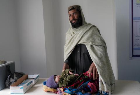 Abdul stands above his baby daughter laying on a table preparing her to recevie an immunisation shot