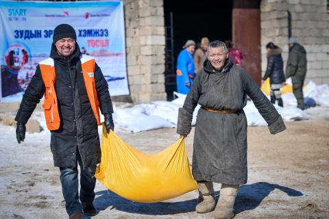 Two men, one wearing an orange World Vision vest, carry a yellow bag of animal fodder