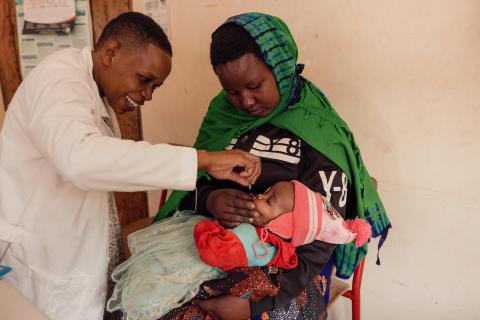 A community health worker in a white lab coat administers an oral vaccine to a baby being held by her mother