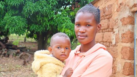 A mother holding a baby next to a stick and mud house.