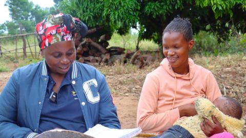 Two women sitting on a grass mat, one holding a baby.