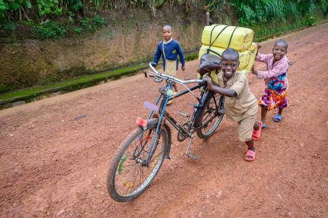 Three children push a bicycle with four yellow water jugs attached the the back.