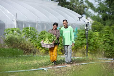A man and woman smile while walking in front of a greenhouse. The woman holds a basket of food.