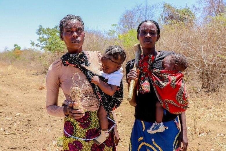 Two women carrying young children look to the camera with serious expressions. The terrain behind them is dry.