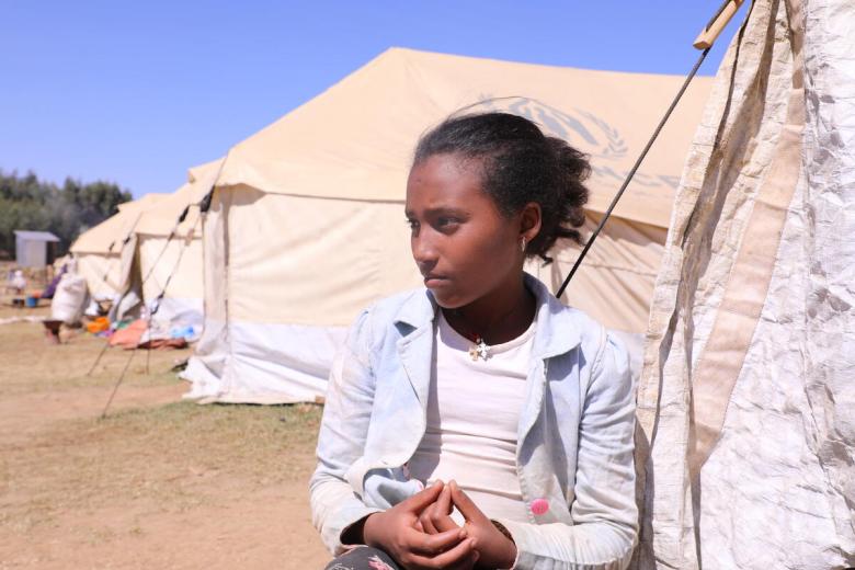 a young girl stands outside the refugee tent she lives in
