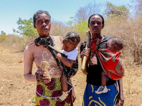 Two Zambian woman, backed by drought affected land, stand with their children looking at the camera.