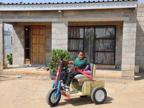 Reithabetse, a 7-year-old girl from Lesotho, smiling outside her home while sitting on her new mobility cart.