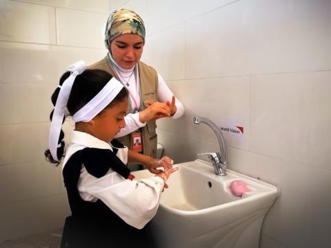 A young girl in a school uniform washes her hands with the help of a female World Vision staff member