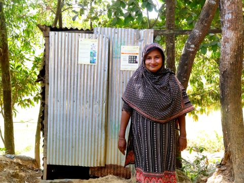 Tafura standing in front of her newly built household toilet in Ramu, Cox’s Bazar, improving her children’s health through safe water, hygiene awareness, and sanitation access.