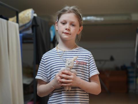 A young girl in a blue and white striped shirts looks into the camera while holding a small, white bunny stuffed animal. 