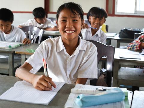 A young girl in a classroom smiles at the camera while sitting at a desk. She holds a pencil over sheets of paper.