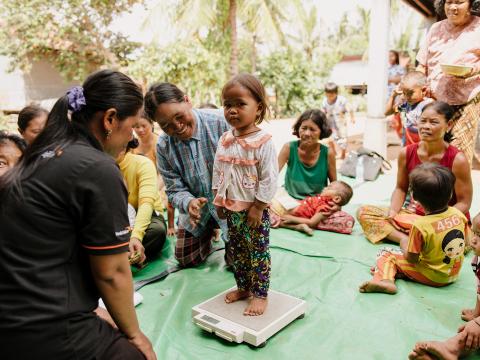 A young Cambodian girl stand on a scale in the middle of a circle of women and children