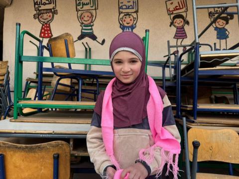 A young girl wearing a head scarf smiles slightly with stacked school desks behind her. 