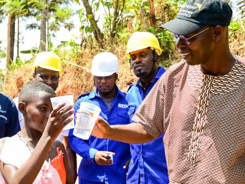 A man wearing a World Vision logoed hat and a young girl touch plastic water cups while 3 engineers watch in the background.
