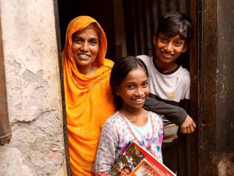 Nargis stands smiling with her young daughter and son in the doorway of their home in a Mirpur slum.