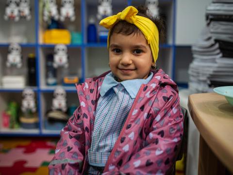 A young girl wearing a blue shirt, pink jacket, and yellow headband, smiles at the camera