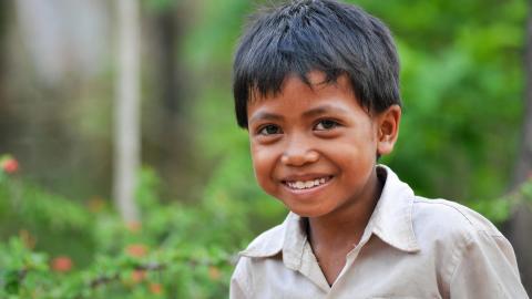 Khmer boy smiles at the camera with lush green foliage behind him.