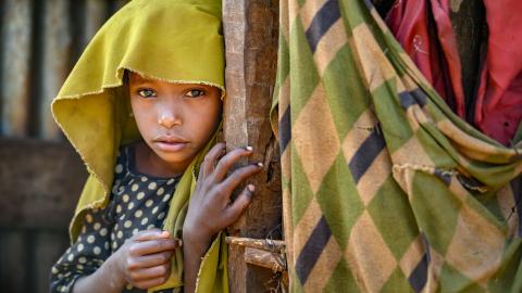 A young girl peeks shyly from behind a wooden post, eyes wide with wonder.