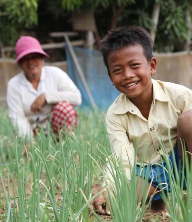 Khmer boy in the foreground kneeling in a grassy field with his mum in the background