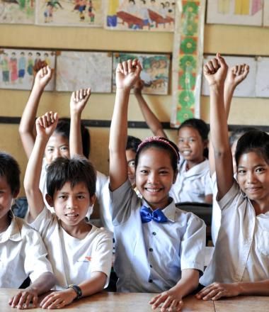 Group of Khmer students smiling with their hands raised in the air sitting at desks inside a classroom