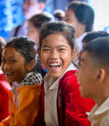 Group of Khmer children sitting on the floor of a classroom with one ecstatic girl looking directly at the camera