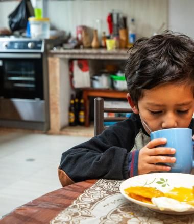 young boy drinks out of a cup at a kitchen table and a plate of food sits in front of him