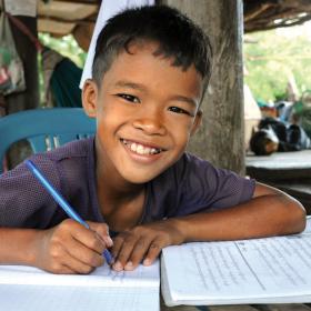 Khmer boy smiles at the camera with pen in hand, writing in his school books, Cambodia.
