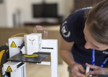 Woman looks as a 3D printer prints using yellow plastic